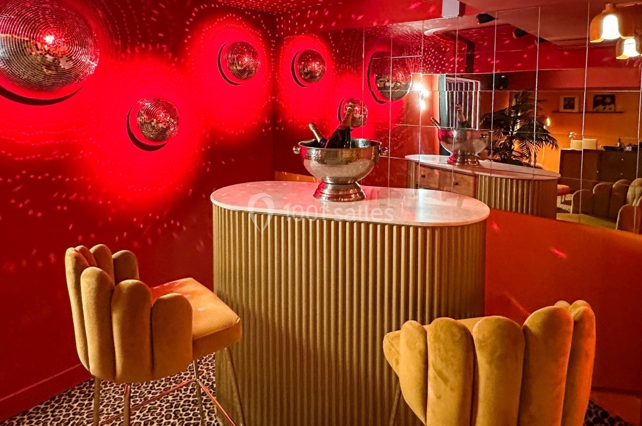 Bar intimiste avec murs rouges, table ovale en marbre, tabourets jaunes et éclairage tamisé reflété par des miroirs.