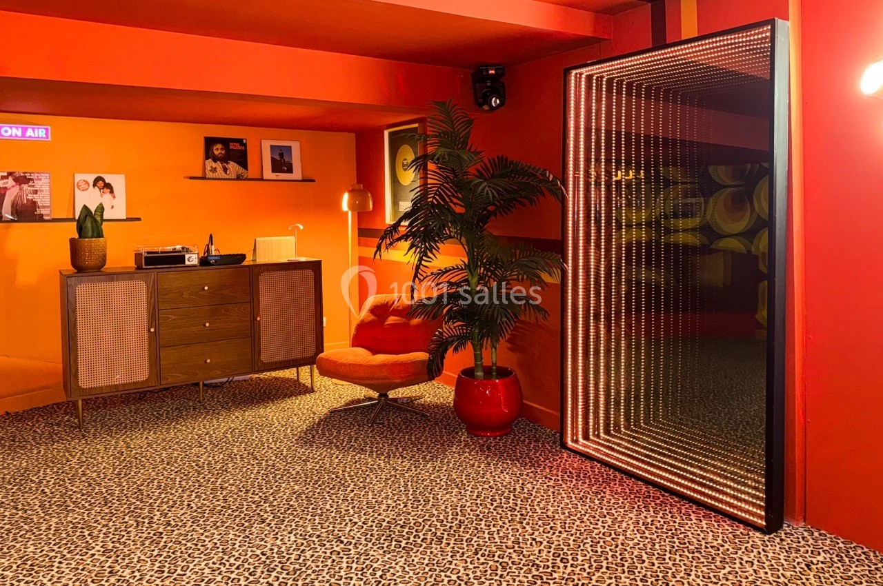 Salon rétro avec murs orange, tapis léopard, mobilier vintage, plante en pot et miroir lumineux vertical.