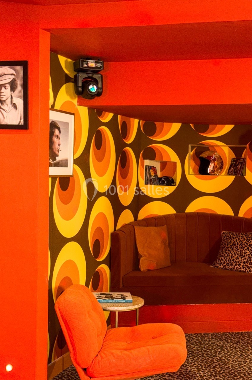 Salon rétro avec fauteuil orange, banquette marron, papier peint à motifs circulaires orange et jaune, et photos au mur.