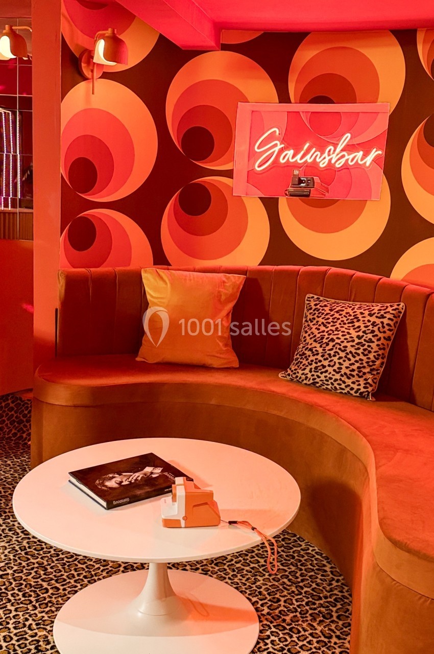 Banquette en velours orange avec coussins, table basse blanche, décor rétro avec motifs circulaires et néon ’Salonbar’.