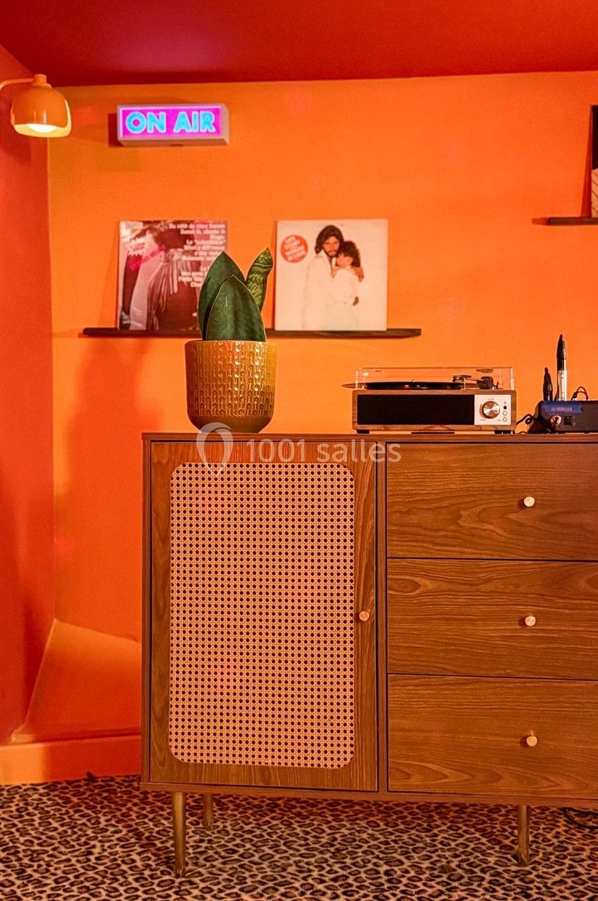 Meuble en bois avec porte en cannage, plante en pot, platine vinyle et disques sur des étagères, mur orange en arrière-plan.