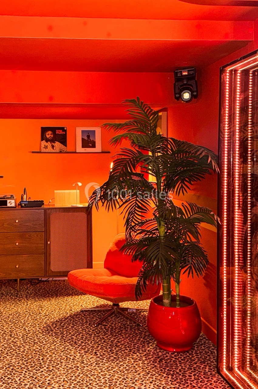 Intérieur rétro avec fauteuil rouge, plante en pot, meuble en bois et murs orange vif, éclairage chaleureux.