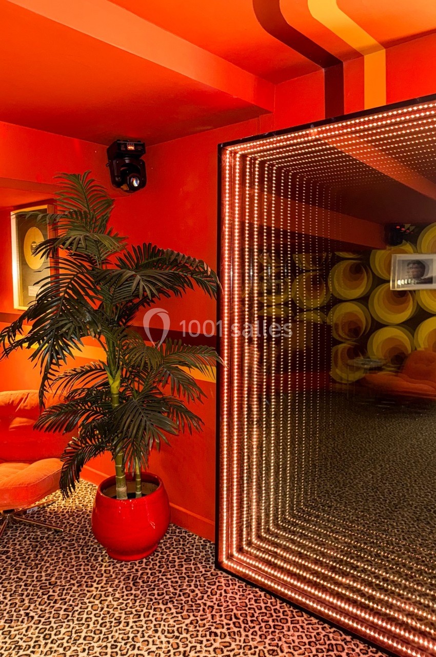 Salle au décor rétro avec murs rouges, fauteuil orange, plante en pot rouge et miroir lumineux à effet infini.