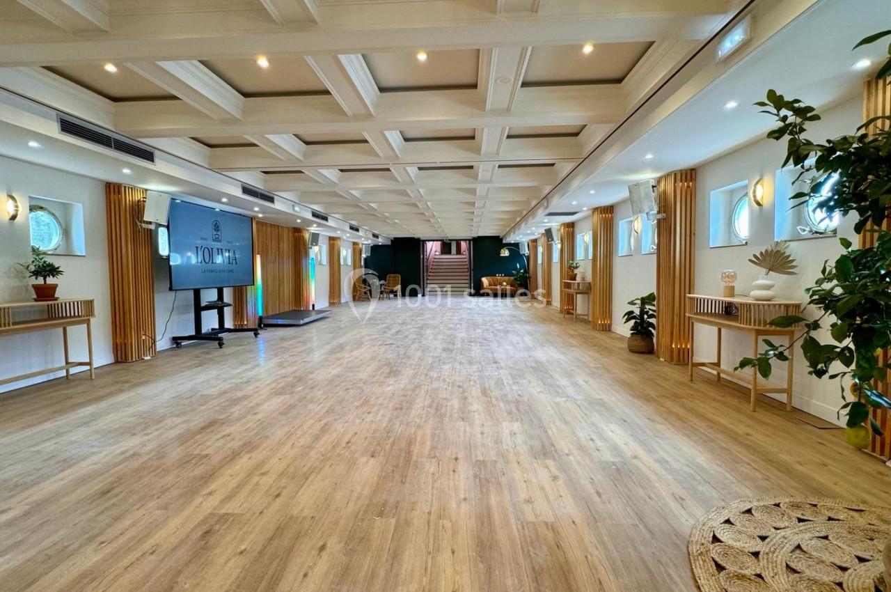 Salle lumineuse avec parquet clair, plantes décoratives, mobilier en bois et écran, éclairée par des hublots et des spots.