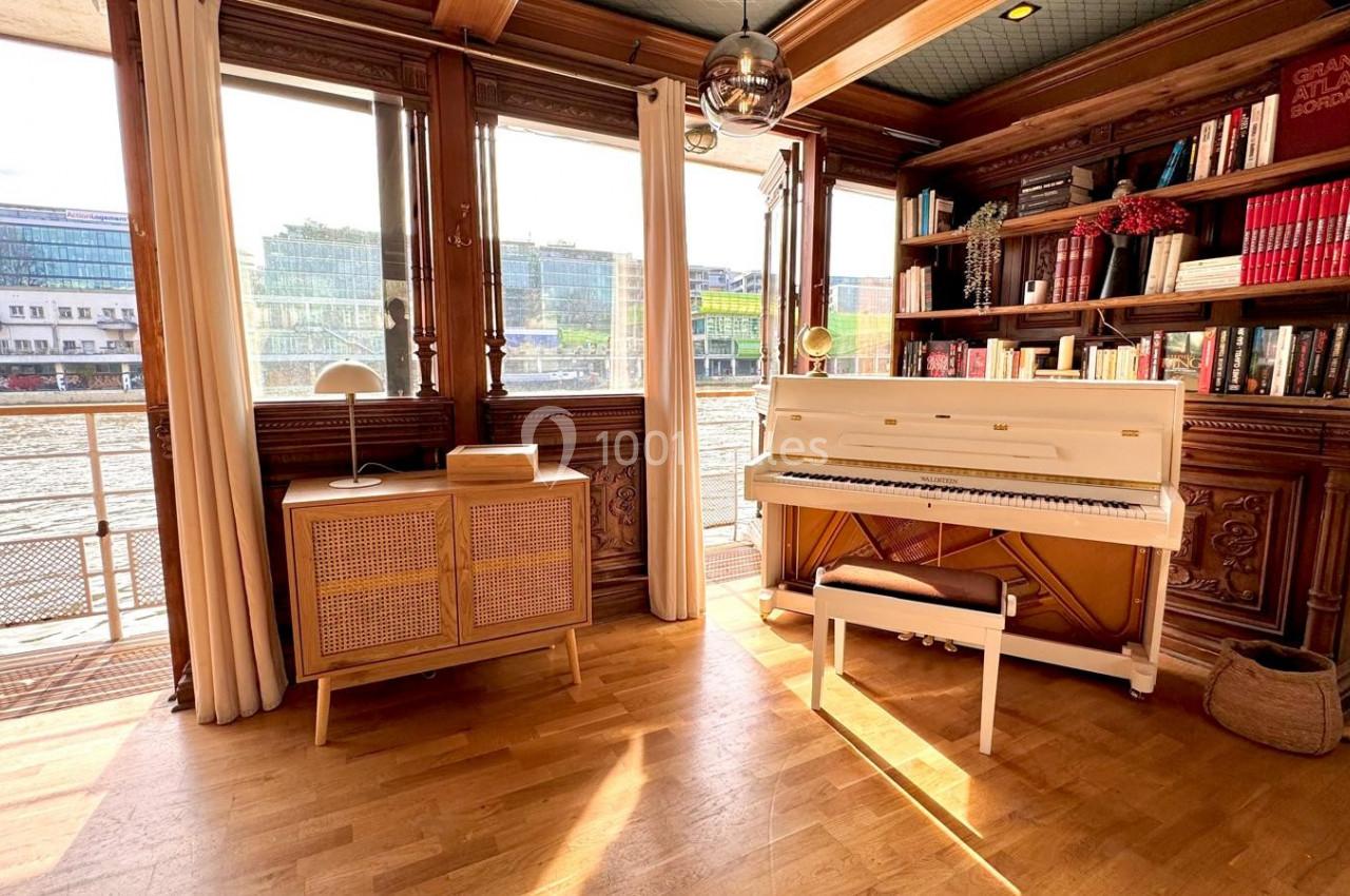 Salon lumineux avec parquet, piano blanc, bibliothèque en bois, et vue sur un canal à travers de grandes baies vitrées.