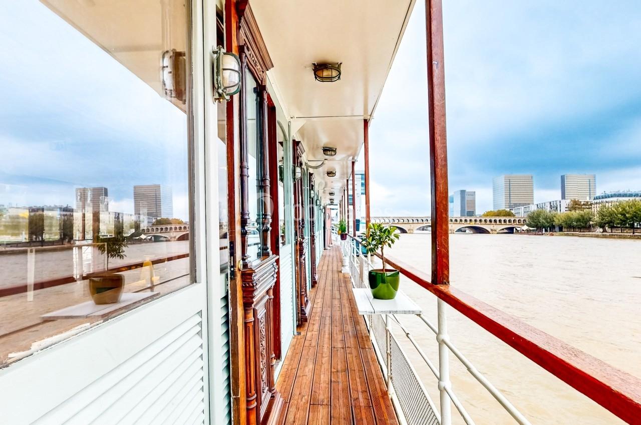 Vue d'un ponton en bois longeant une péniche avec des bâtiments modernes et un fleuve en arrière-plan.