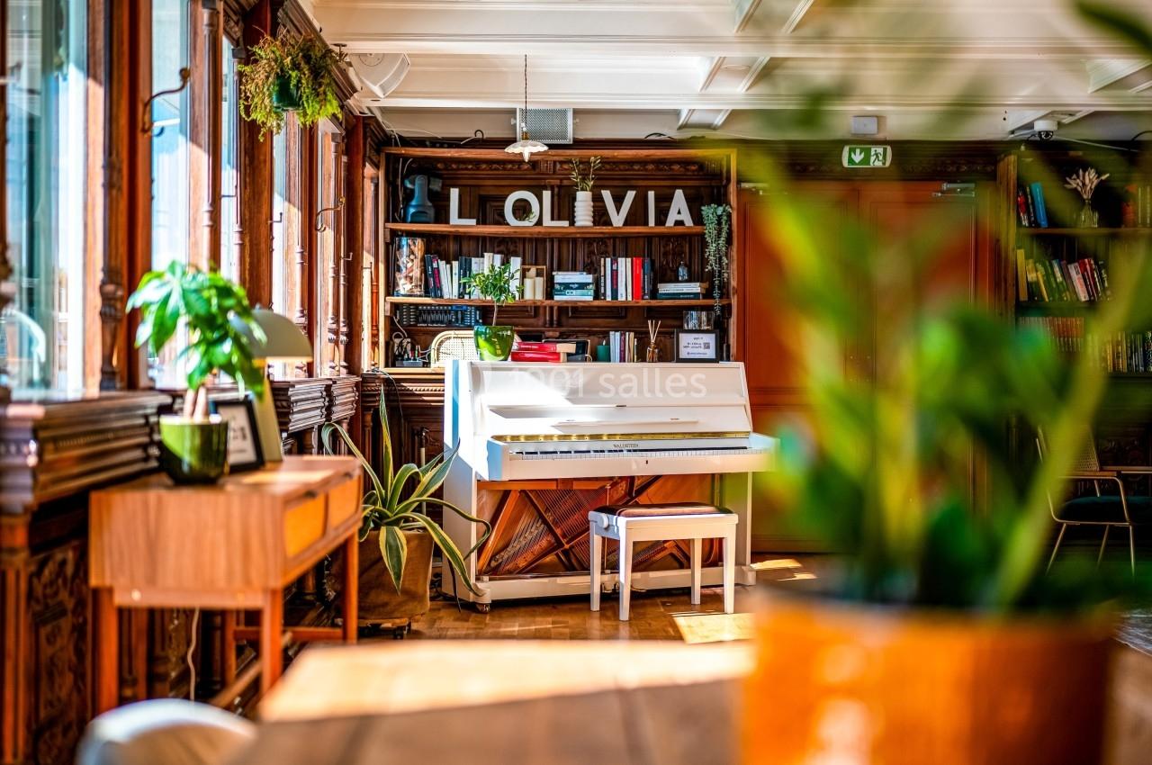 Piano blanc dans une pièce lumineuse avec des plantes, des étagères en bois et des livres en arrière-plan.