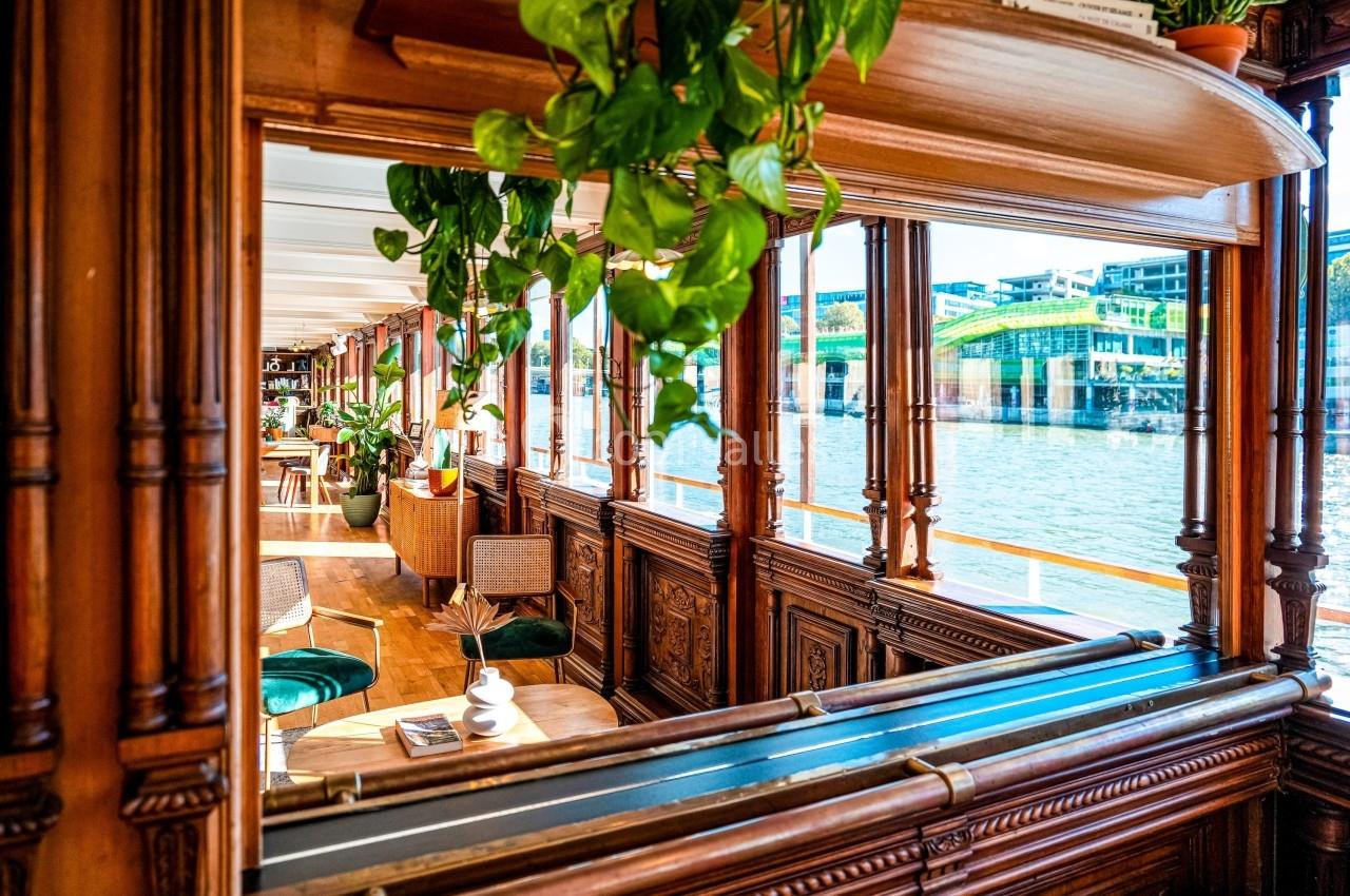 Intérieur d'une péniche en bois avec des plantes suspendues, des fauteuils et une vue sur un fleuve et des bâtiments…