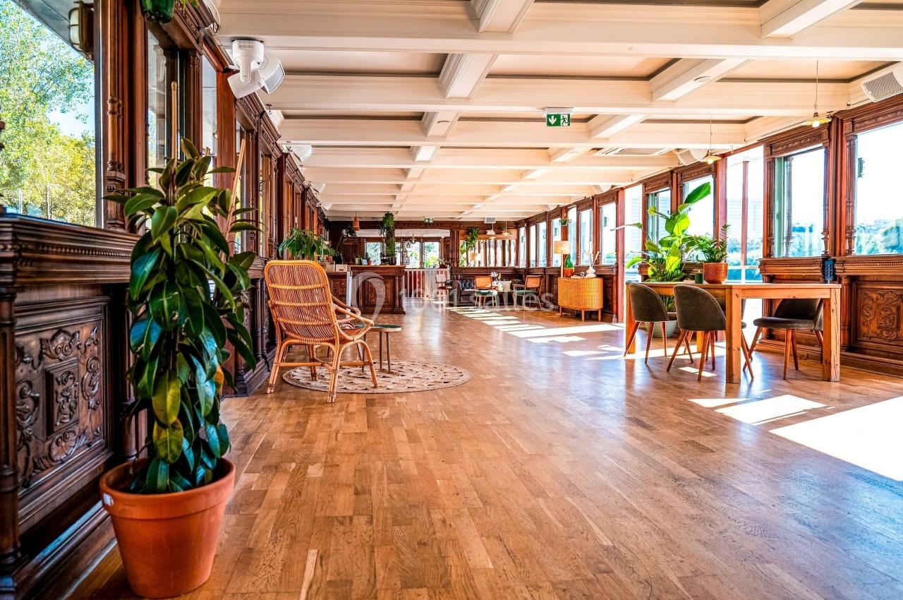 Salle lumineuse avec parquet en bois, grandes fenêtres, plantes en pot et mobilier en rotin et bois.