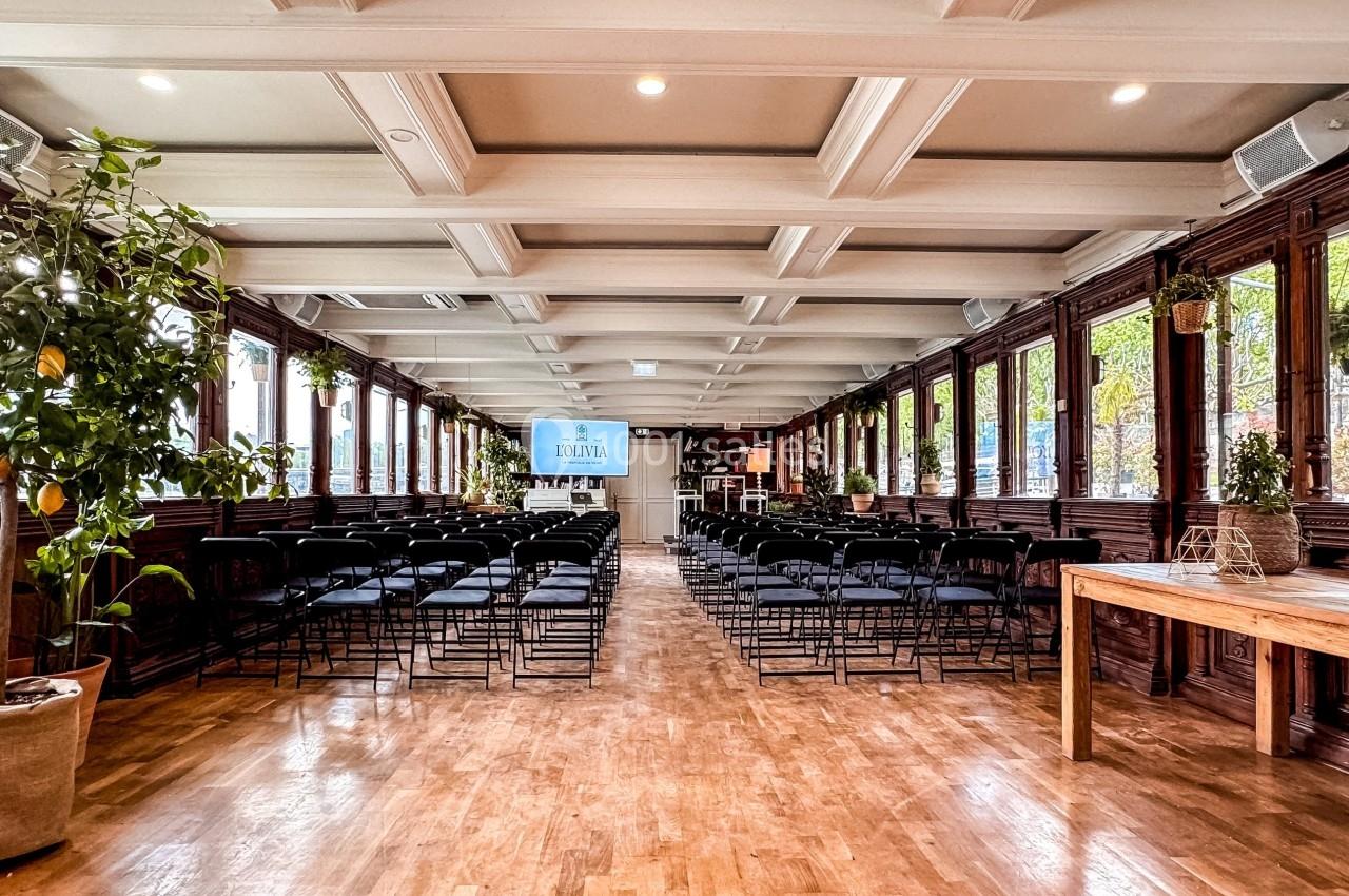 Salle lumineuse avec parquet, rangées de chaises noires, plantes en pot et grandes fenêtres donnant sur l'extérieur.