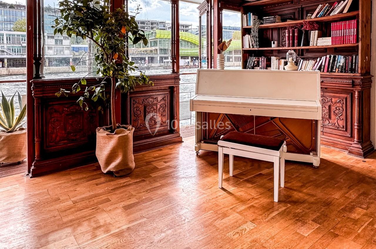 Piano blanc avec tabouret dans une pièce lumineuse, entouré de plantes, d'étagères en bois et d'une vue sur l'extérieur.
