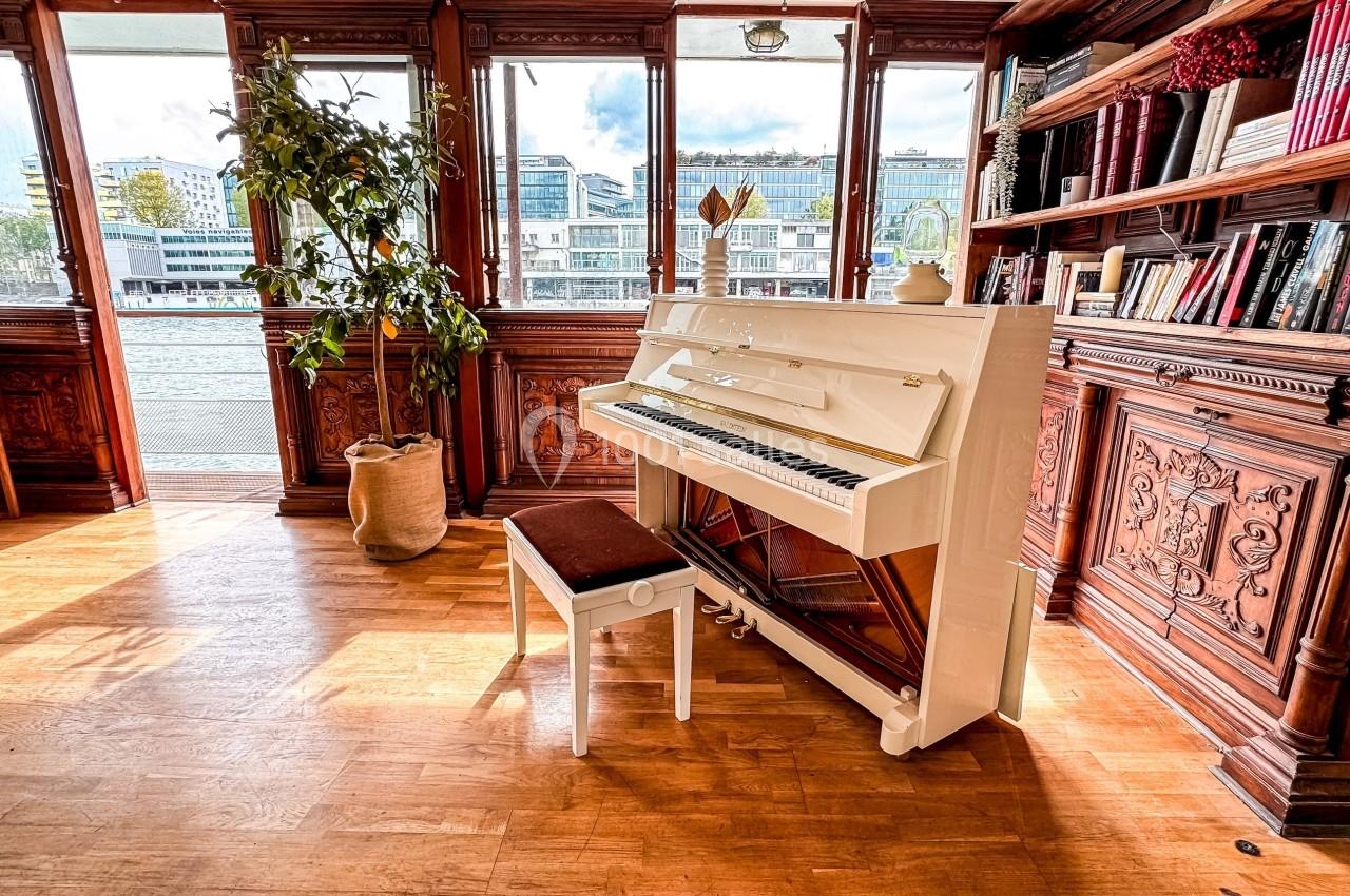 Piano droit blanc avec tabouret, placé dans une pièce lumineuse avec parquet, bibliothèque en bois et vue sur des bâtiments.