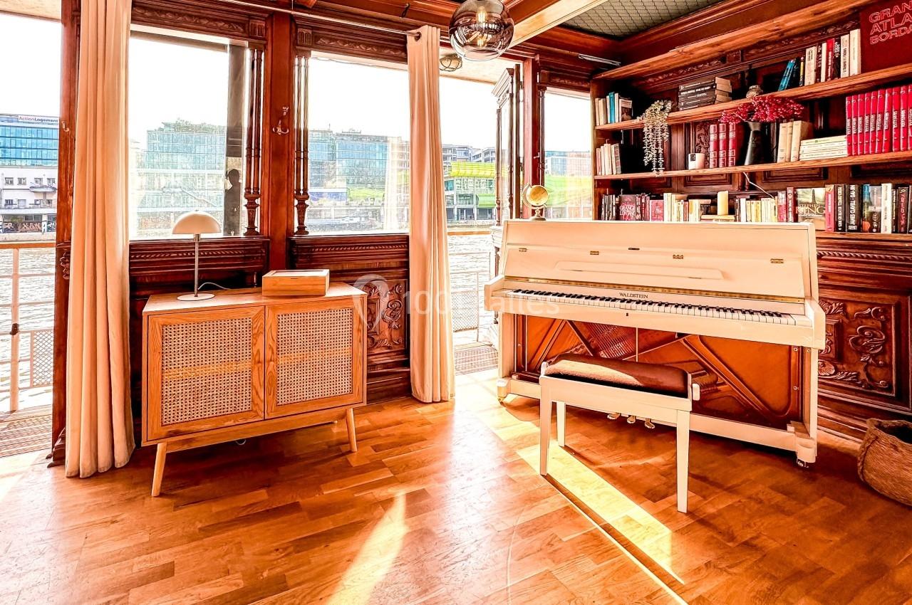 Intérieur lumineux avec un piano blanc, une bibliothèque en bois, et une vue sur l'eau à travers de grandes fenêtres.