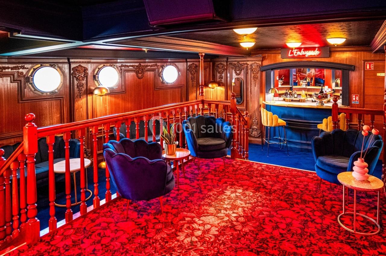 Salon chaleureux avec fauteuils bleus, tapis rouge à motifs, boiseries sculptées et bar éclairé en arrière-plan.