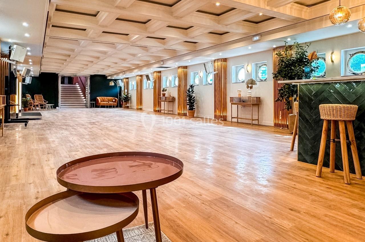 Salle spacieuse avec parquet clair, mobilier moderne, bar vert et éclairage chaleureux, décorée de plantes et de hublots.