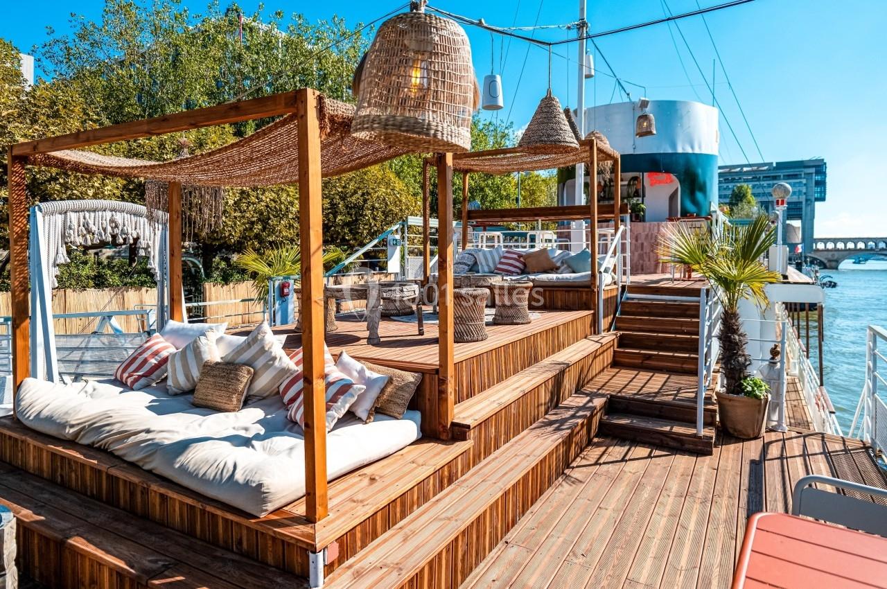 Terrasse en bois avec lits de repos, coussins et parasols en osier, située au bord d'une rivière sous un ciel dégagé.