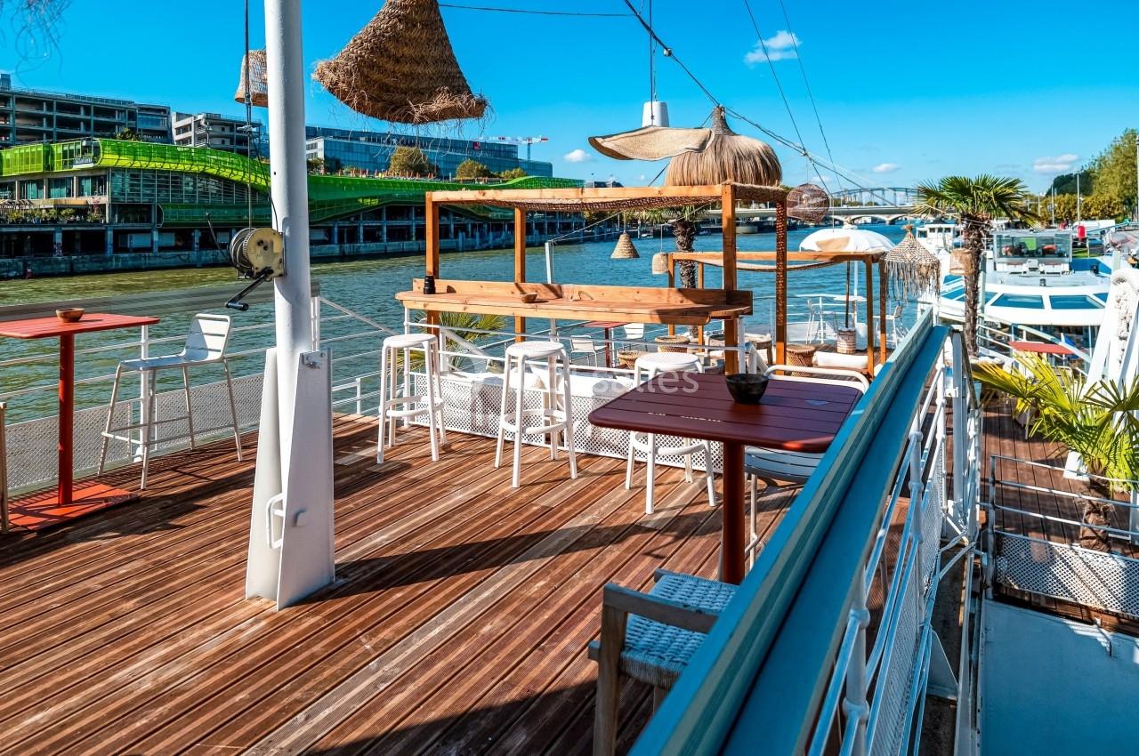 Terrasse en bois avec tables et chaises sur une péniche, bordée par un fleuve et des bâtiments modernes en arrière-plan.