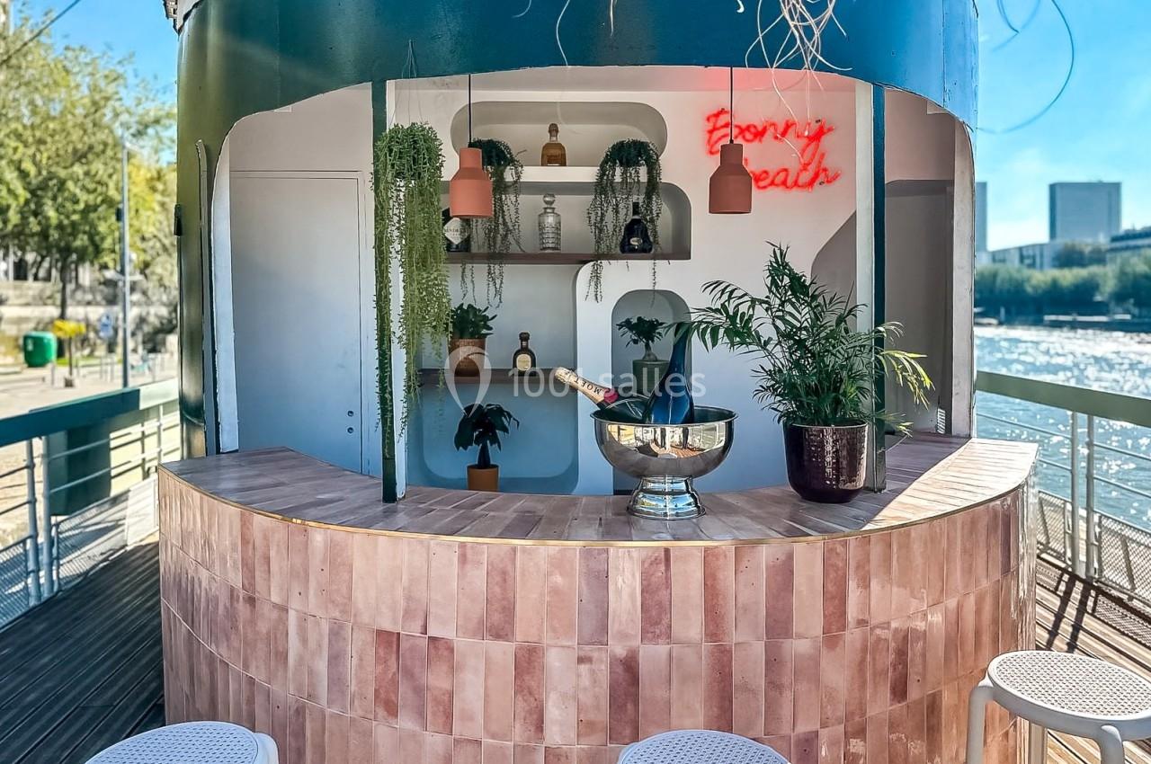 Bar extérieur avec comptoir en carreaux roses, plantes décoratives, seau à champagne et enseigne lumineuse rouge.