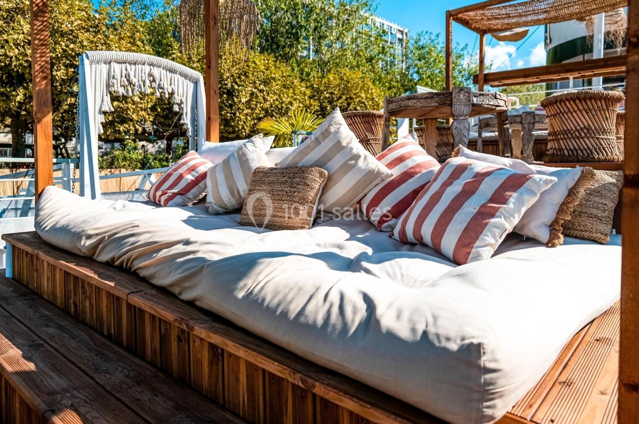Lit de repos extérieur avec coussins rayés et un auvent en bois, entouré de végétation et baigné de lumière naturelle.