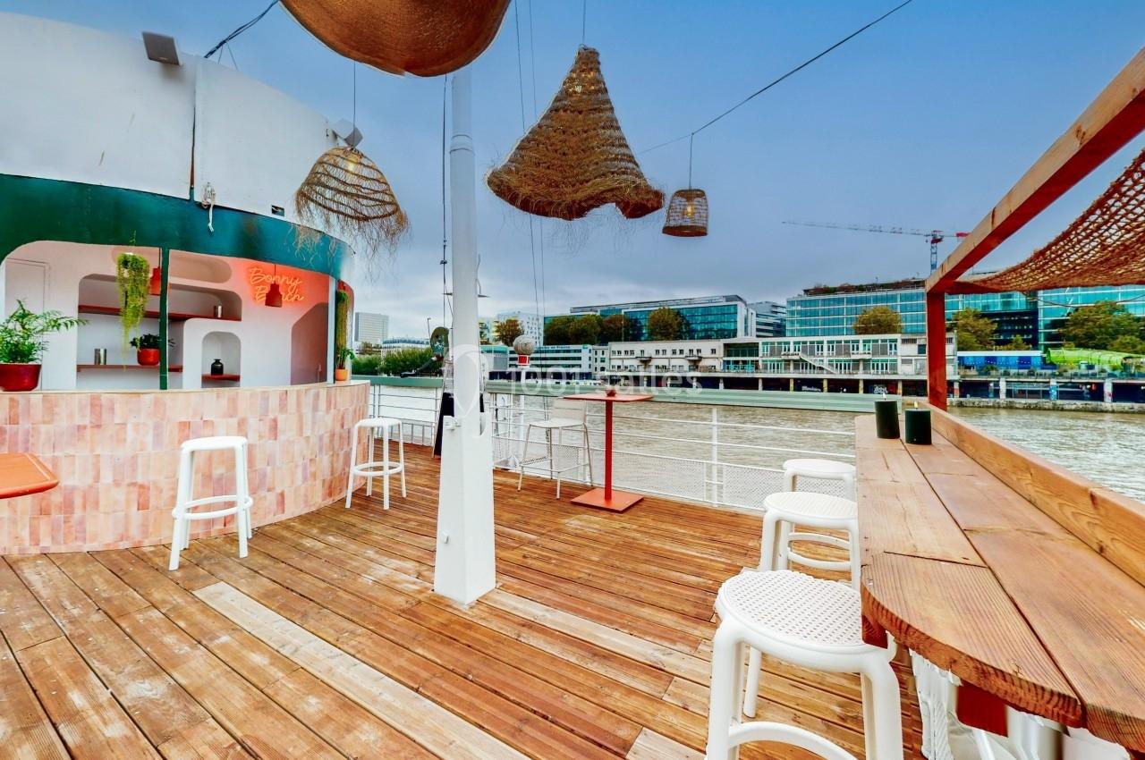 Terrasse en bois avec bar, tabourets et suspensions décoratives, donnant sur une rivière et des bâtiments modernes.