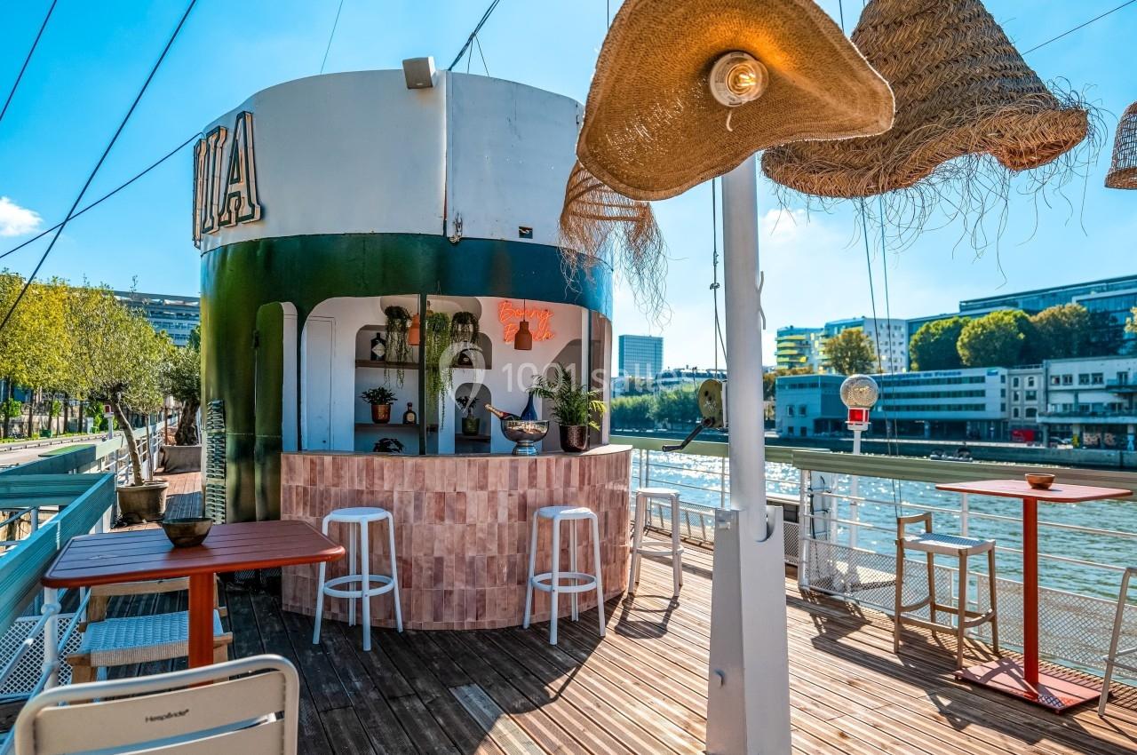 Bar extérieur sur une péniche, avec tabourets, tables et décorations en osier, au bord d'une rivière ensoleillée.