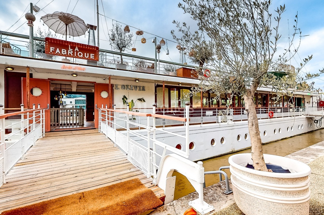 Entrée d'un restaurant sur une péniche avec terrasse, accessible par une passerelle en bois bordée de rambardes blanches.