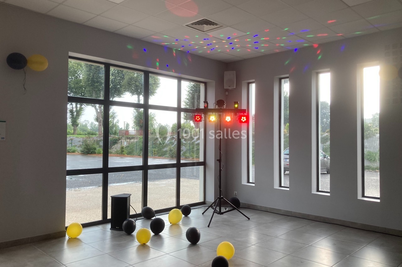 Salle lumineuse avec des ballons noirs et jaunes au sol, éclairage coloré et projecteurs près des fenêtres.