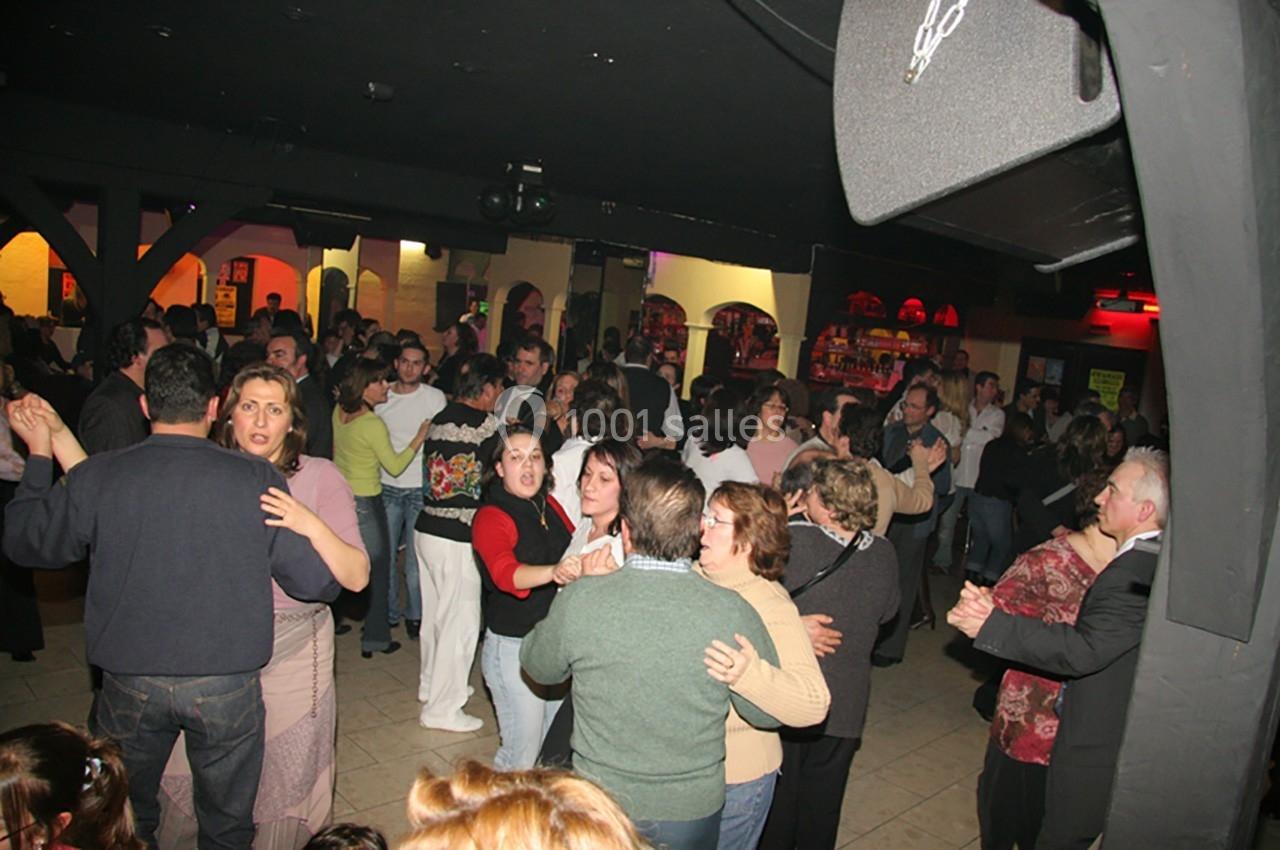 Des personnes dansent en couple dans une salle animée avec un éclairage tamisé et un bar en arrière-plan.