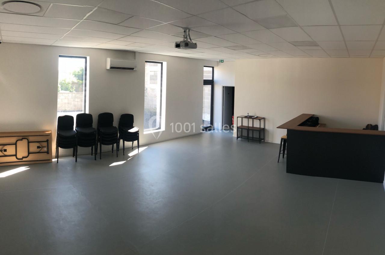 Salle vide avec sol gris, murs blancs, chaises empilées, meuble en bois, bar noir et lumière naturelle provenant des…
