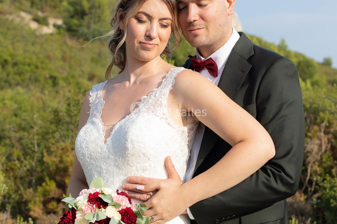 Un couple en tenue de mariage pose dans un paysage naturel verdoyant, la mariée tenant un bouquet de fleurs.