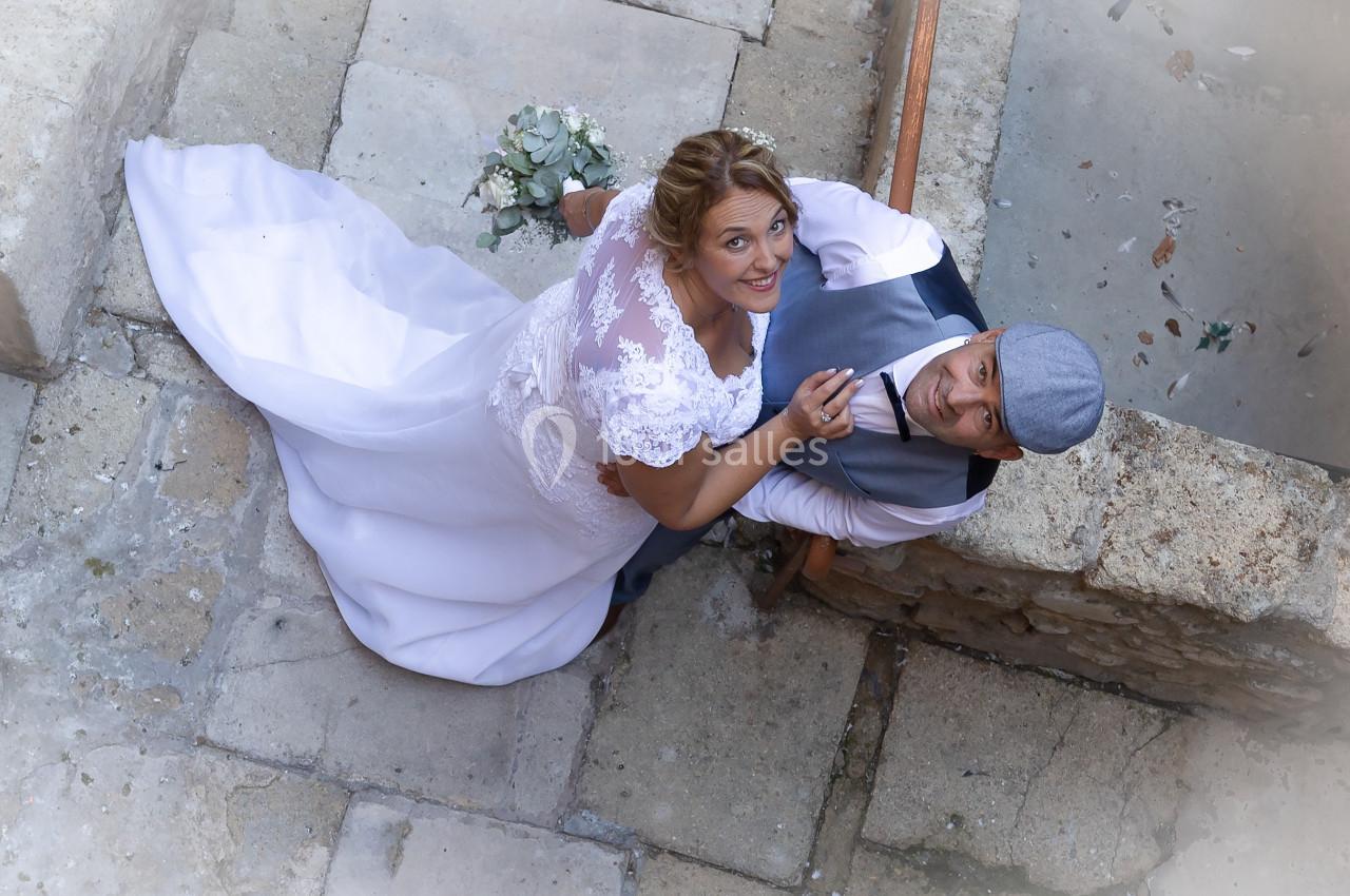 Un couple de mariés vu de dessus, souriant, la mariée en robe blanche tenant un bouquet.