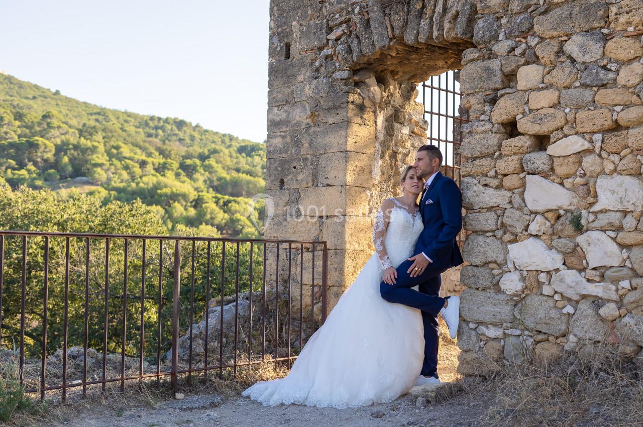 Un couple en tenue de mariage pose près d'un mur en pierre avec une vue sur un paysage verdoyant en arrière-plan.