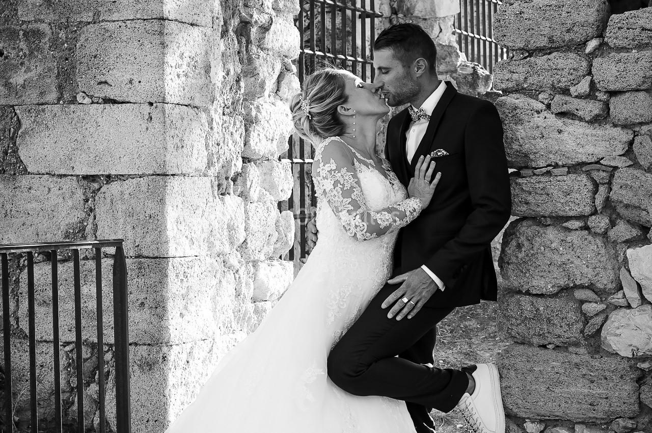 Un couple de mariés s'embrasse tendrement devant un mur en pierre dans un cadre extérieur ancien.