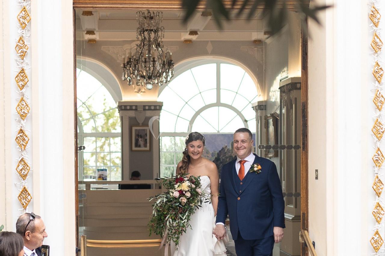 Un couple de mariés souriants sort d'un bâtiment élégant sous le regard des invités.