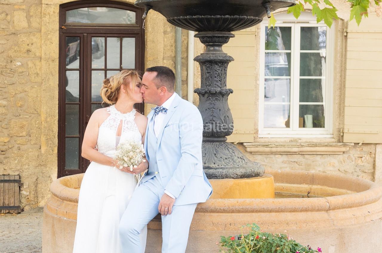 Un couple en tenue de mariage pose près d'une fontaine devant un bâtiment en pierre avec des fenêtres.