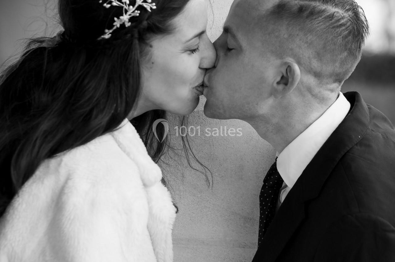 Un couple s'embrasse tendrement près d'une colonne, la femme porte une couronne ornée et un manteau clair.
