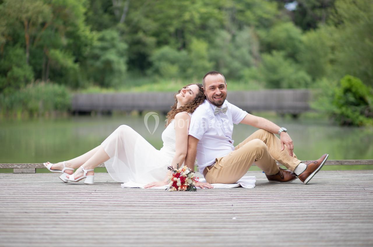 Un couple assis dos à dos sur un pont en bois, près d'un étang entouré de verdure.