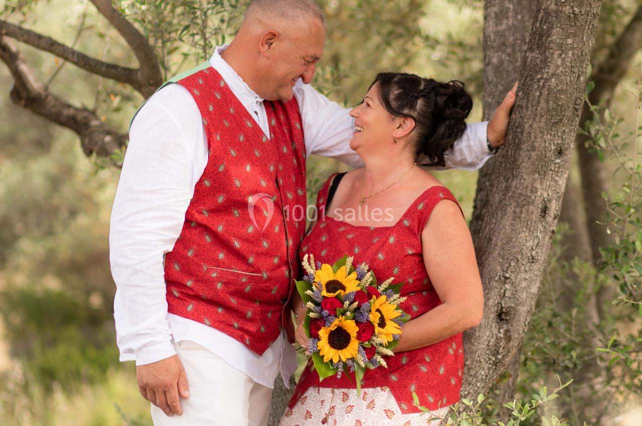 Un couple souriant en tenue estivale, l'homme appuyé contre un arbre, la femme tenant un bouquet de tournesols.