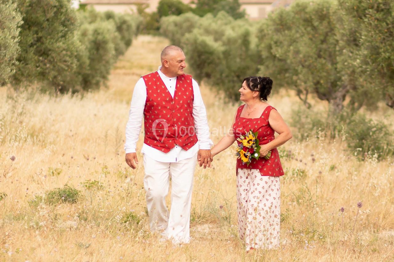 Un couple en tenue assortie rouge et blanche marche main dans la main dans un champ bordé d'oliviers.