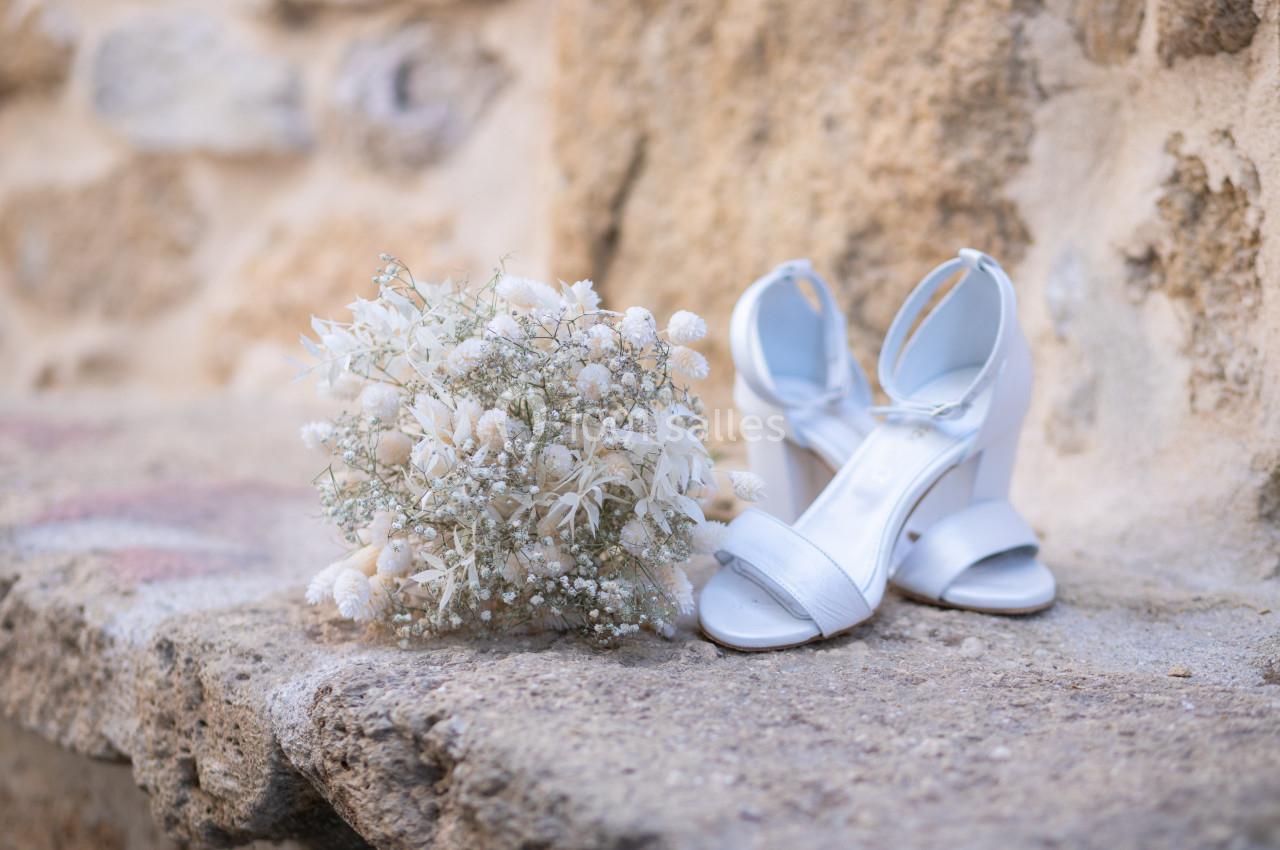 Bouquet de fleurs séchées blanches et sandales blanches à talons posés sur une surface en pierre beige.