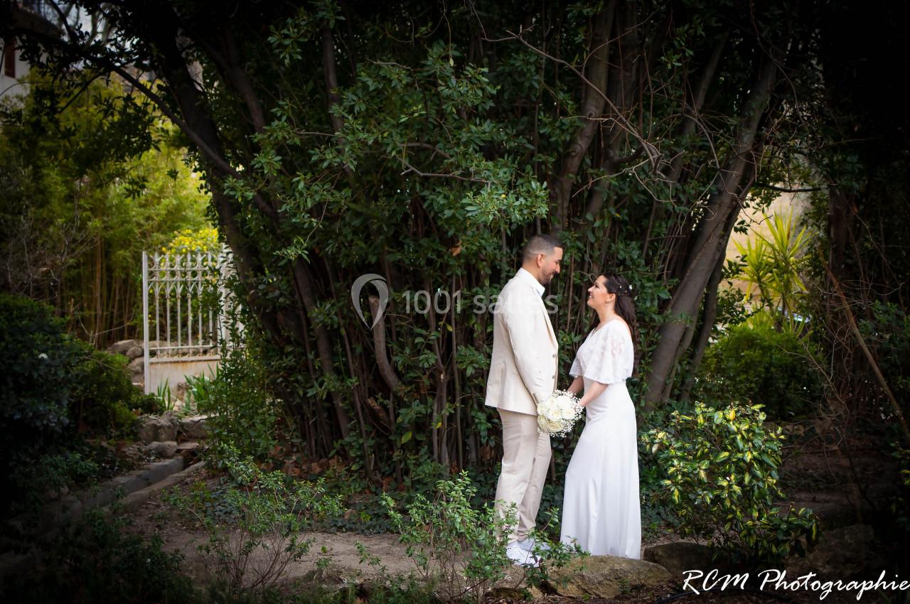 Un couple en tenue de mariage se tient face à face devant un grand arbre dans un jardin verdoyant.