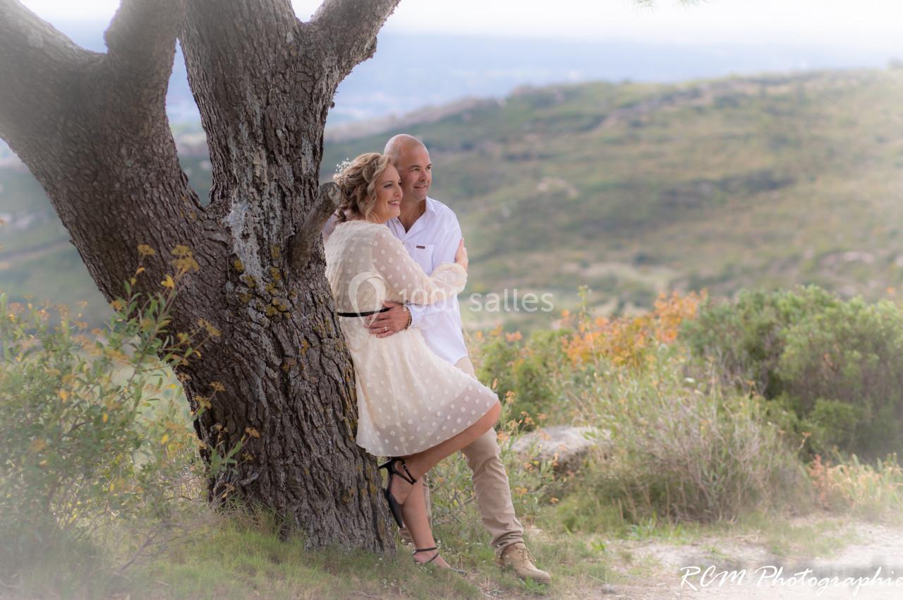 Un couple souriant s'appuie contre un arbre dans un paysage naturel vallonné.