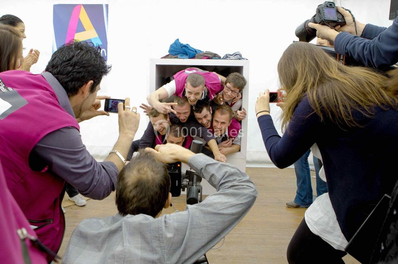 Des personnes empilées dans une boîte rient et posent, entourées de photographes capturant la scène.