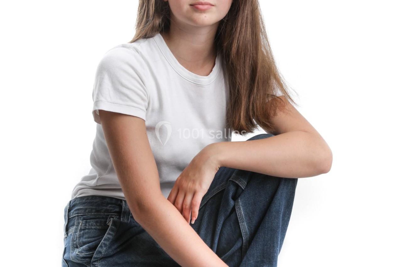 Jeune femme assise sur le sol, portant un t-shirt blanc et un jean, posant devant un fond blanc.