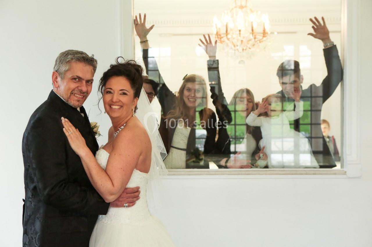 Un couple en tenue de mariage pose devant une fenêtre où des invités souriants font des gestes joyeux.