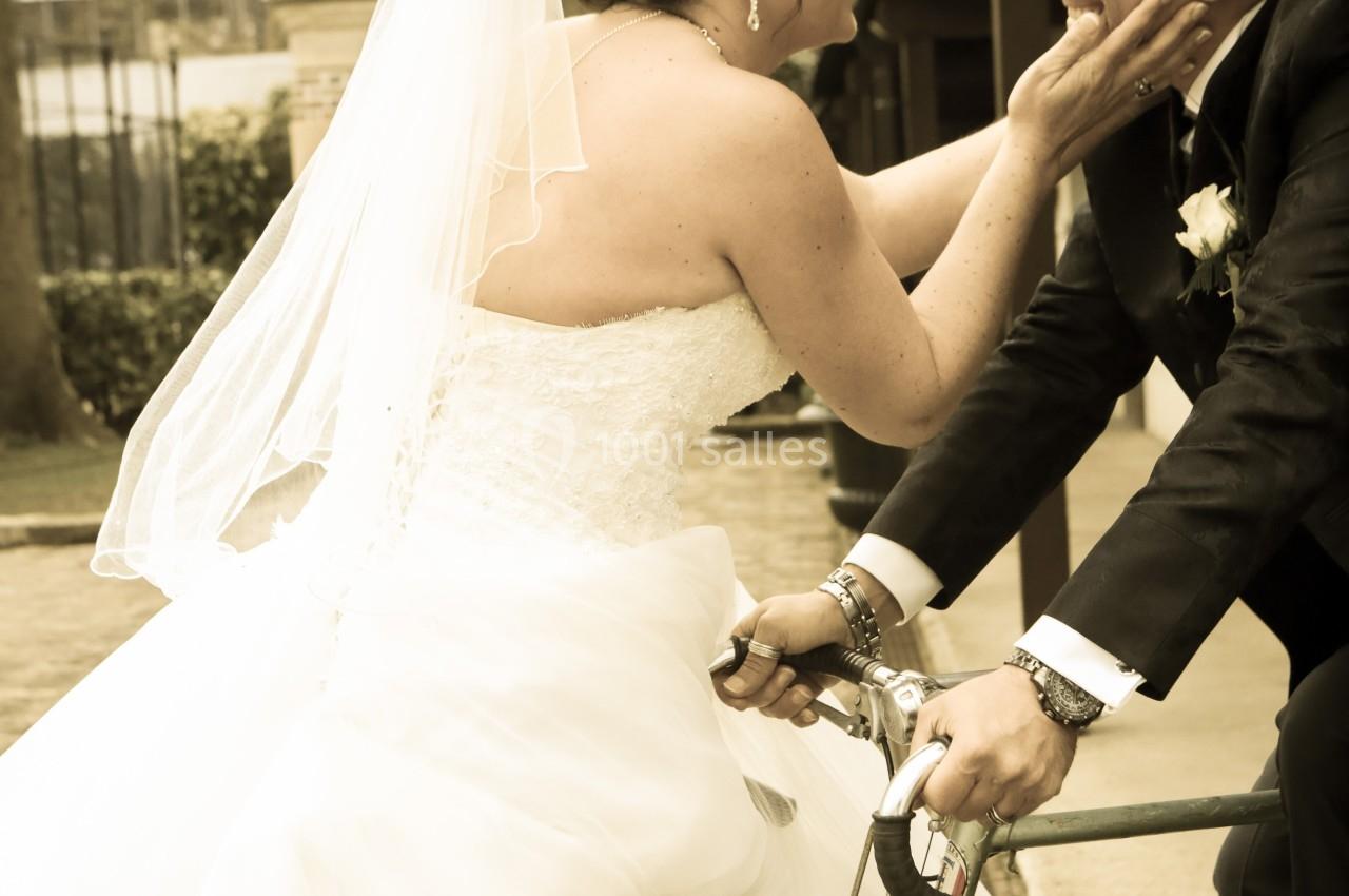 Une mariée souriante touche le visage du marié assis sur un vieux vélo, dans une ambiance extérieure chaleureuse.