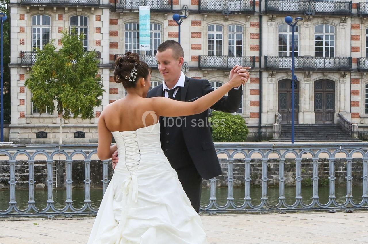 Un couple en tenue de mariage danse sur une place pavée devant un bâtiment ancien au bord de l'eau.