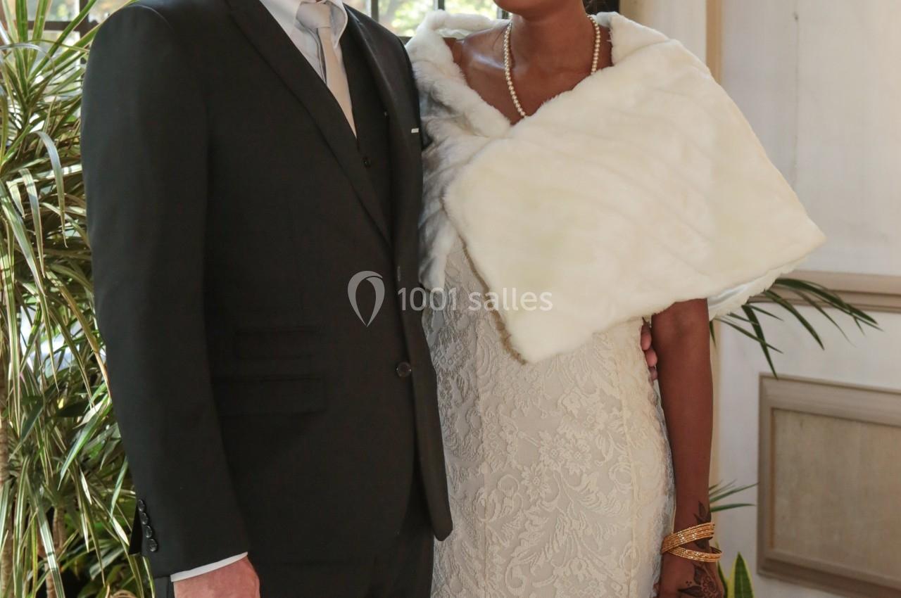 Un couple en tenue de mariage pose devant une fenêtre avec des plantes en arrière-plan.