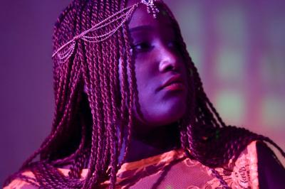 Femme souriante portant un blazer imprimé coloré, un t-shirt gris et un jean, posant devant un fond blanc.