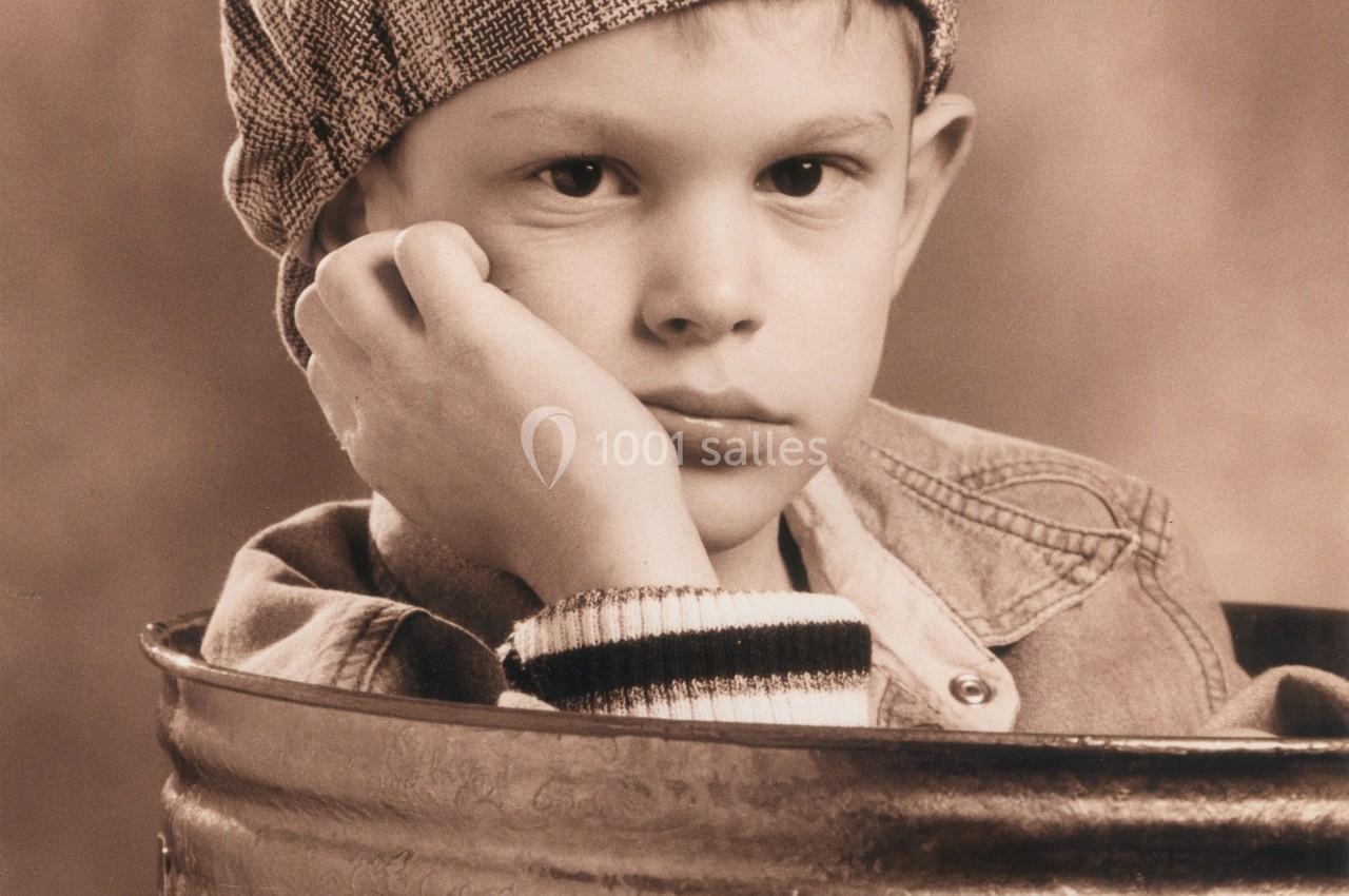 Un enfant pensif portant une casquette, appuyé sur une poubelle métallique dans un décor vintage.